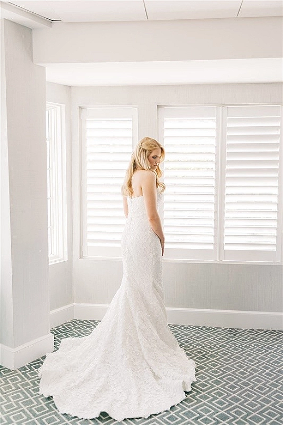 Bridal portrait of a blonde bride in a strapless lace wedding dress with a long train, looking over her shoulder by white shutters indoors