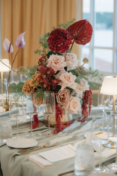 Reception centerpiece with a wedding floral centerpiece of roses, orchids, and greenery beside place settings and a glowing table lamp by a window