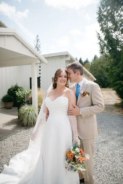 Couple portrait of bride holding bouquet as groom hugs her, lace veil flowing beside a white barn on a cloudy day outdoors