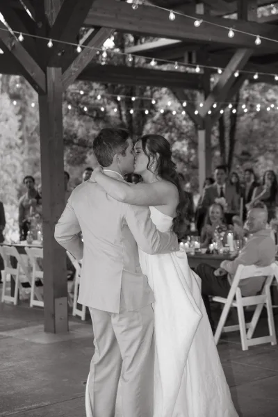 First dance as bride in a wedding gown and groom in a suit kiss under string lights in a wood pavilion with guests watching