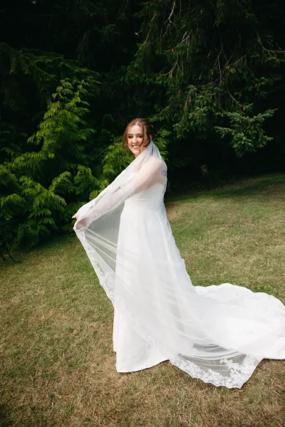 Bridal portrait of a smiling bride in a wedding dress holding a long veil with lace trim on a grass lawn before evergreen trees