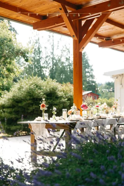 Reception tablescape on an outdoor reception table with bud vase florals, candles, lace runner, and string lights under a wood pergola