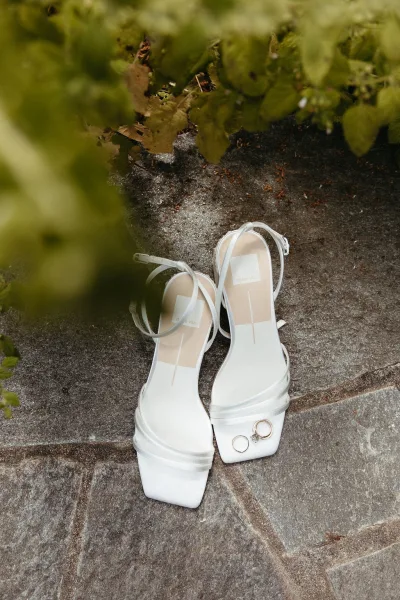 Wedding shoes with bridal shoes styling, white strappy heels topped with wedding rings on stone pavement beside green foliage