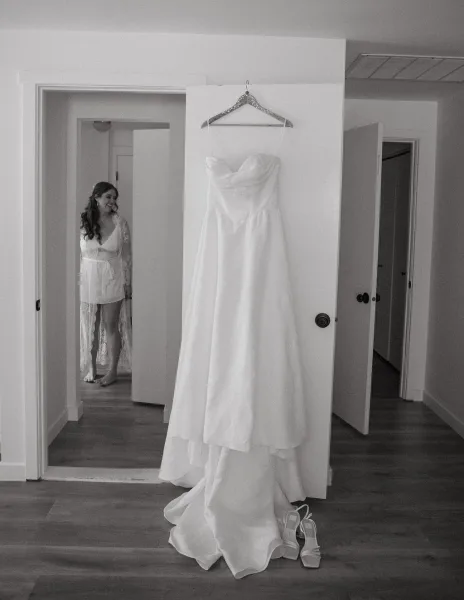 Wedding dress hanging on a doorway hanger, a strapless bridal gown with white heels below in a bright bedroom hallway