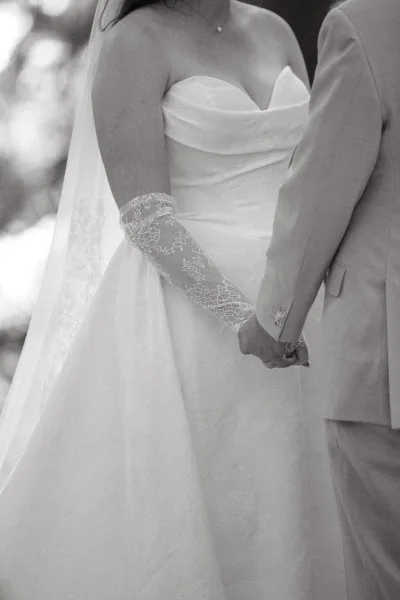 Couple portrait of bride and groom holding hands, lace gloves on a strapless gown and veil, outdoors with softly blurred trees behind