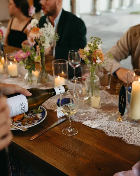 Reception tablescape with wedding table decor, lace runner, taper and votive candles, wildflower bud vases, and place cards on a wooden table indoors