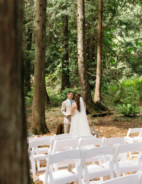 Ceremony moment at an outdoor forest wedding ceremony with bride in veil and groom in tan suit exchanging vows under string lights amid ferns