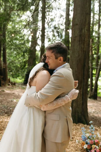 Wedding hug as bride and groom embracing on a tree-lined forest path under string lights, her veil and lace gloves visible beside wildflowers