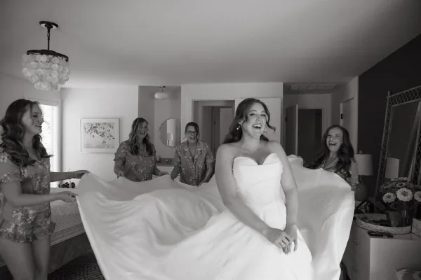 Bride getting ready as bridesmaids help with her strapless wedding dress, bouquet in hand, in a chandelier-lit bedroom by window blinds