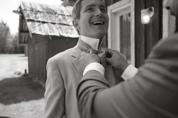 Groom getting ready as groomsmen adjust his tie and boutonniere on a light gray suit by a rustic barn doorway opening to the yard