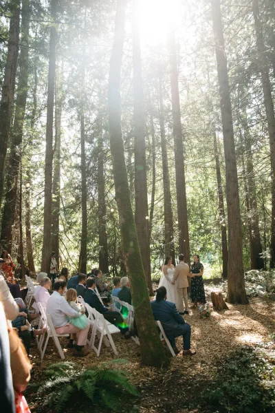 Wedding ceremony with guests seated on white folding chairs beneath string lights in a sunlit forest aisle lined with florals