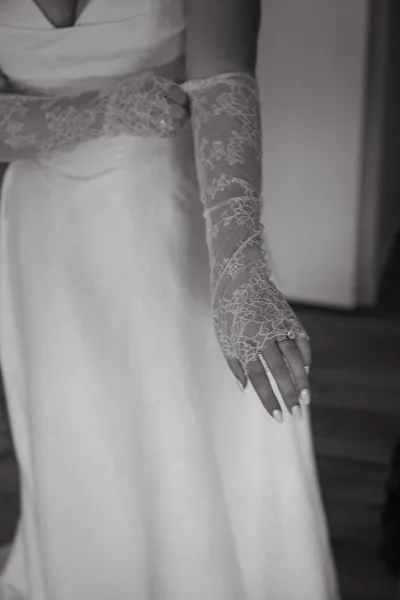 Bridal gloves in sheer lace wedding gloves style, paired with a wedding dress and engagement ring, shown indoors against a plain wall
