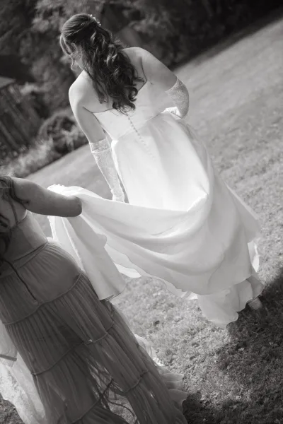 Bride walking from behind holding her wedding dress bustle, wearing lace gloves and a hair comb on a tree-lined lawn outdoors