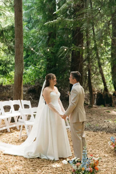 Ceremony moment as bride and groom hold hands on a wood chip aisle under forest trees, veil and reserved chairs with string lights behind