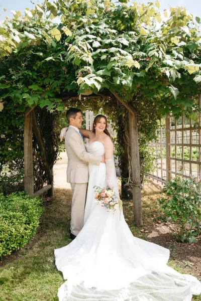 Couple portrait of bride and groom hugging under a vine-covered pergola, bride holding a pastel bouquet and long train, groom in beige suit