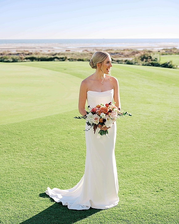 Bridal portrait of a bride in a strapless wedding dress holding a rose-and-greenery bouquet, standing on a grassy lawn by the ocean shore.