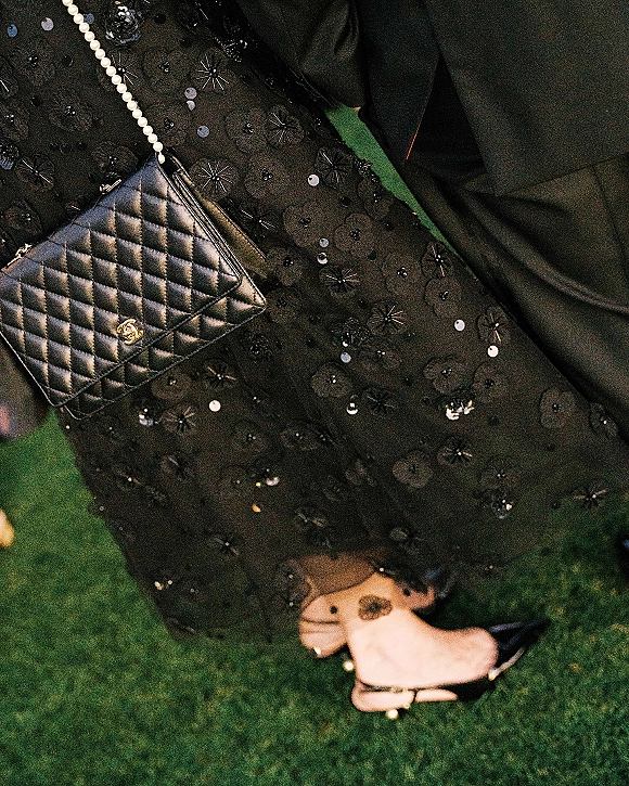 Wedding guest outfit featuring a black wedding guest dress with floral sequins, pearl-strap quilted bag, and slingback heels on a lawn