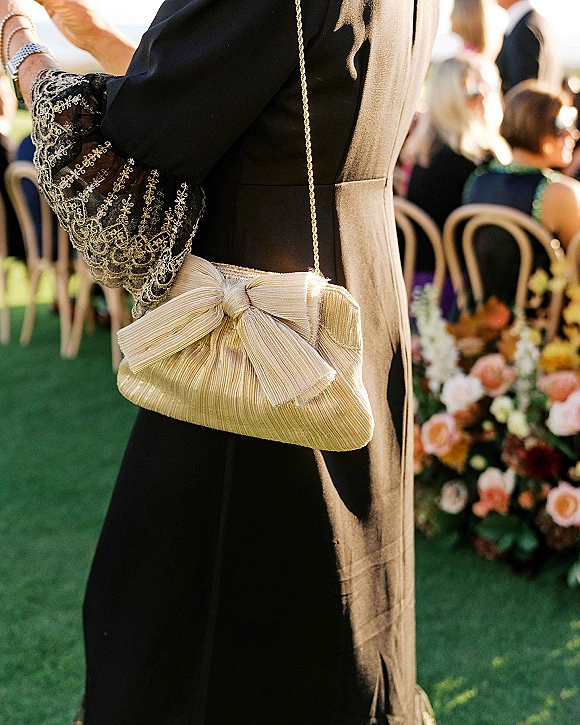 Wedding guest outfit in a black tie dress with embroidered lace sleeve, gold chain clutch and bracelet at an outdoor lawn ceremony