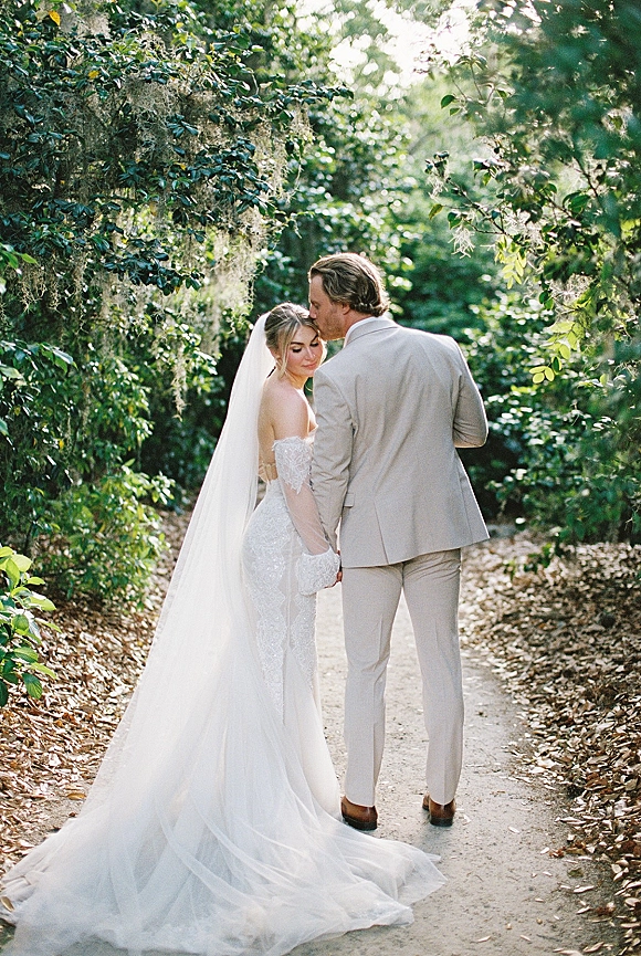 Couple portrait of bride and groom holding hands, walking away on a garden path under Spanish moss, her long veil and lace train flowing