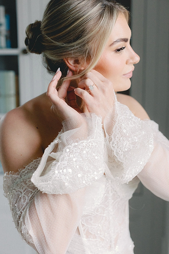 Bridal portrait of a bride getting ready, adjusting her earring by window light, wearing an off-the-shoulder lace dress and updo
