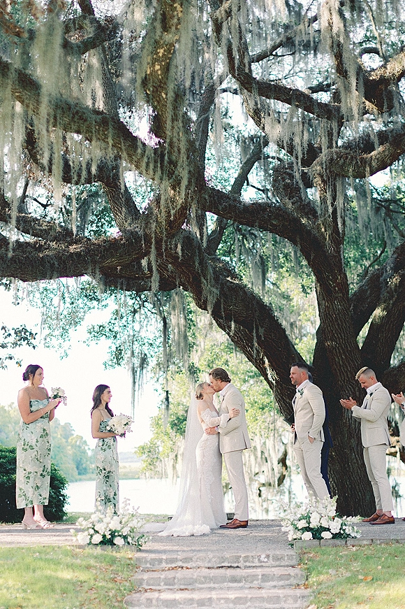 Wedding ceremony kiss as bride and groom embrace under an oak tree with hanging moss, wedding party behind on stone steps by lake