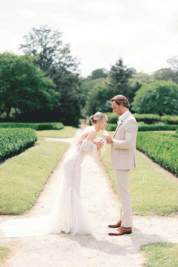 First look moment as bride in strapless lace wedding dress leans toward groom in suit on a garden path under bright sky