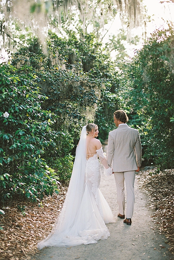 Couple portrait of newlyweds holding hands walking away, bride looking back in long veil and lace dress on a sunlit tree-lined path
