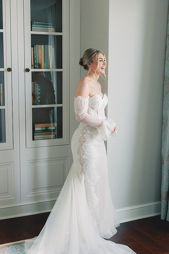 Bridal portrait of a bride laughing in side profile by a window, wearing an off-shoulder lace gown with pearl earrings beside bookshelves