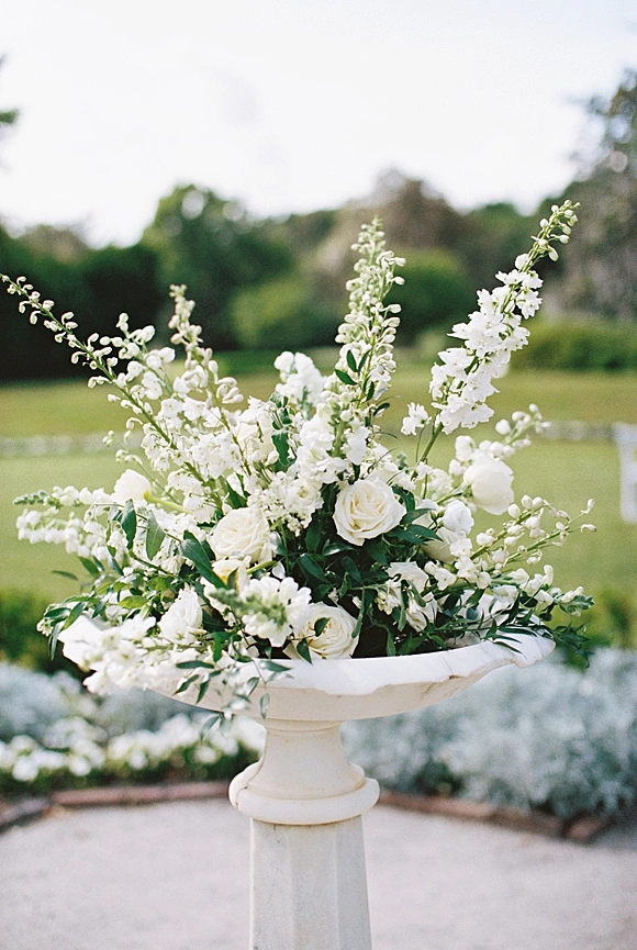 Ceremony floral arrangement in a stone pedestal urn with white roses and greenery beside white chairs on a garden lawn