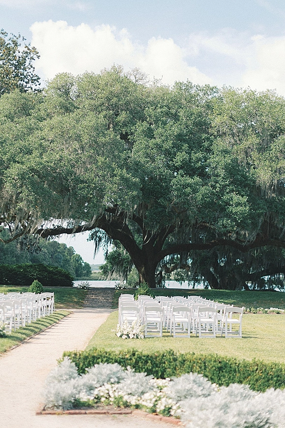 Outdoor ceremony setup with white folding chairs lining a flower-bordered aisle beneath a mossy oak tree by the lake under cloudy sky