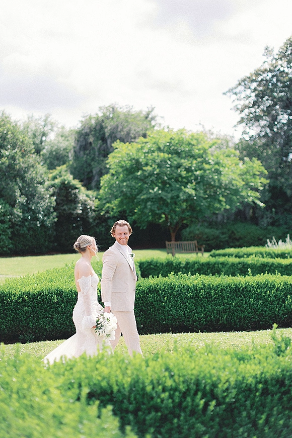 Couple portrait of bride in a lace wedding dress holding a white bouquet beside groom in beige suit in a manicured garden under cloudy sky