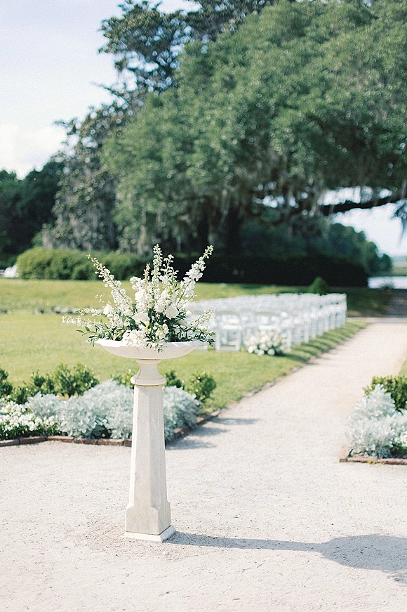 Ceremony setup with white floral urn and greenery accents beside a gravel aisle lined with white flowers and folding chairs on a garden lawn