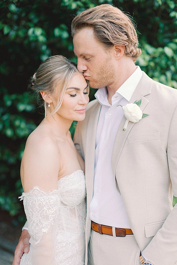 Couple portrait of bride and groom embrace as he kisses her forehead, strapless lace dress and beige suit amid lush garden greenery