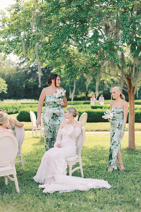 Bride with bridesmaids in a bridal party portrait, bride seated in lace sleeves with long train, bridesmaids holding white bouquets on sunny garden lawn