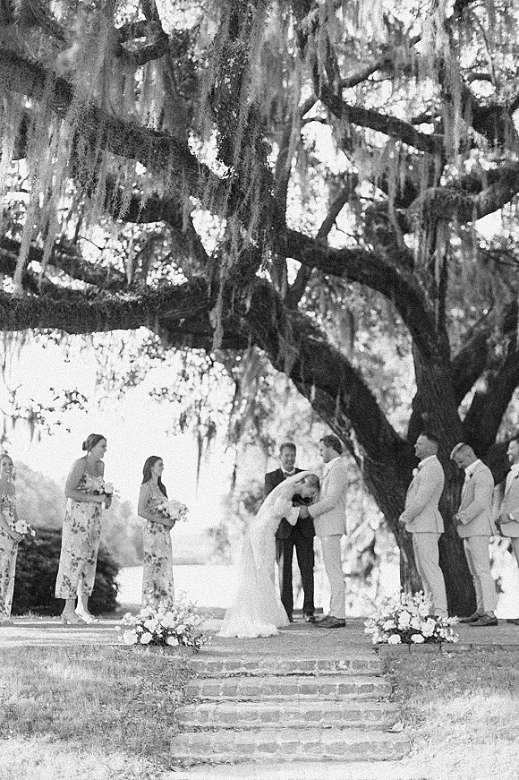 Ceremony moment at an outdoor wedding ceremony as the bride and groom dip kiss beneath a mossy oak tree with bouquets on stone steps