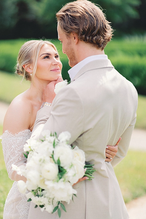 Couple portrait of bride and groom embrace on a green lawn, groom touching her face as she holds a white bouquet with greenery