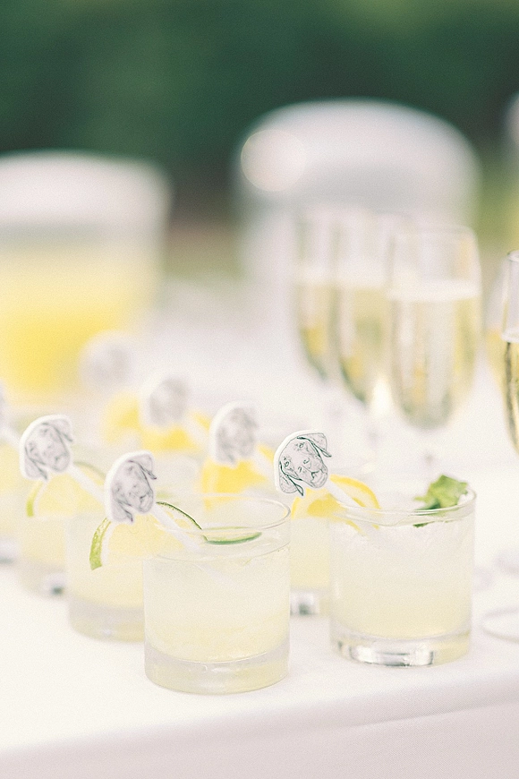 Wedding signature cocktails in clear glasses with lime slices and citrus wedges on a tablecloth, set at an outdoor drink station with greenery