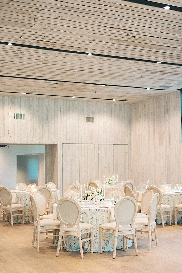 Reception tablescape with round wedding tables dressed in floral tablecloths, white floral centerpieces, candles, and upholstered chairs in a whitewashed wood room