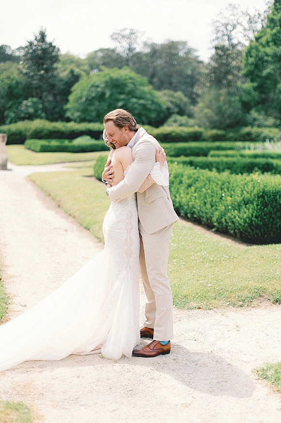 Couple portrait of bride and groom hug on a gravel garden path, her strapless lace gown with long train beside his beige suit