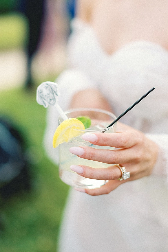 Wedding cocktail in a highball glass with black straw, ice, lemon wedge and lime, held by a manicured hand with engagement ring outdoors