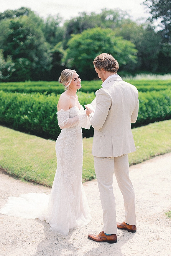 Wedding vows as groom in a beige suit reads from vow cards to bride in an off-shoulder lace gown on a garden gravel path