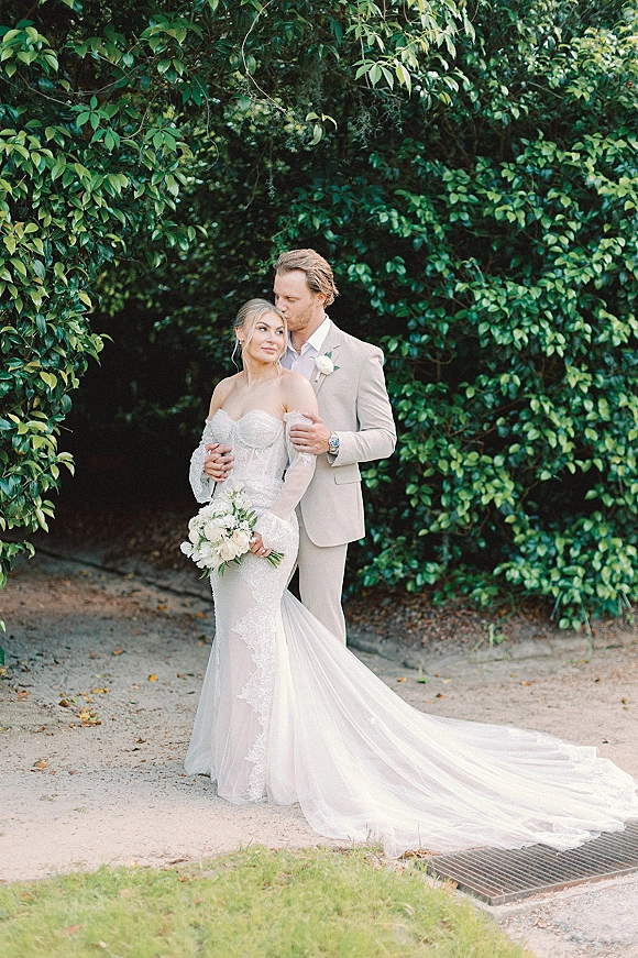 Couple portrait of groom kissing bride’s forehead as she holds a white rose bouquet, lace off-shoulder gown and beige suit by green hedge