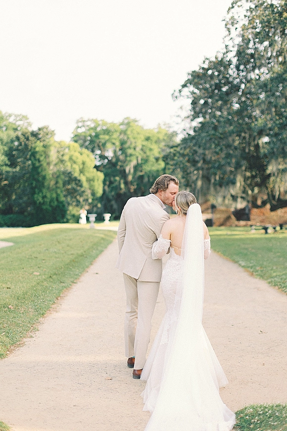 Wedding kiss portrait of bride and groom kissing from behind, her long veil flowing over an off-shoulder dress on a tree-lined path in a park