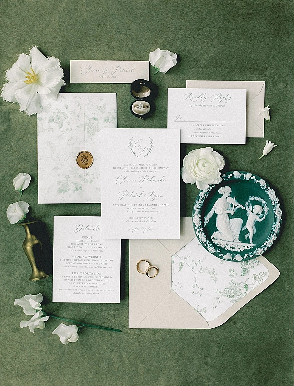 Invitation suite wedding invitation flat lay with wax seal, RSVP and details cards, gold rings, and white flowers on green fabric backdrop
