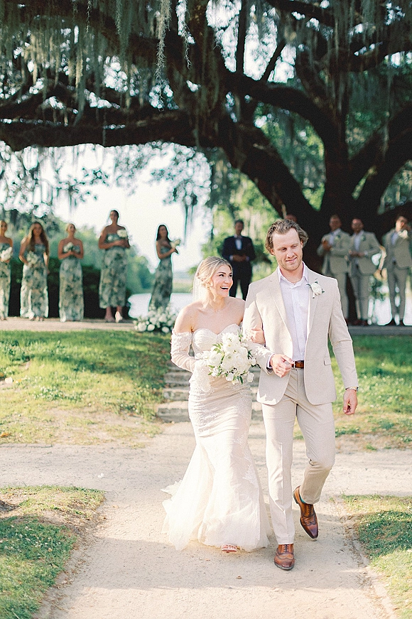 Wedding recessional with bride and groom walking arm in arm, bride holding a white bouquet under oak trees with Spanish moss by water view