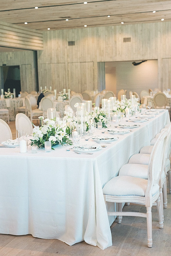 Reception tablescape with long head table decor featuring white floral garlands, greenery, taper candles and glassware in a wood-paneled room