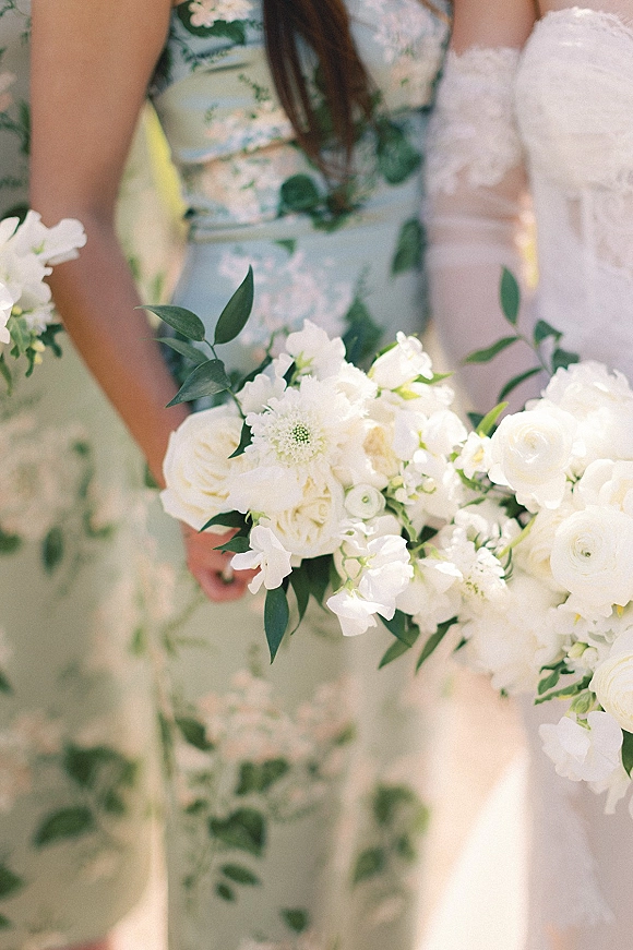 Bridesmaid bouquet of white blooms and greenery held beside a floral print dress and strapless wedding gown in sunlit outdoor greenery