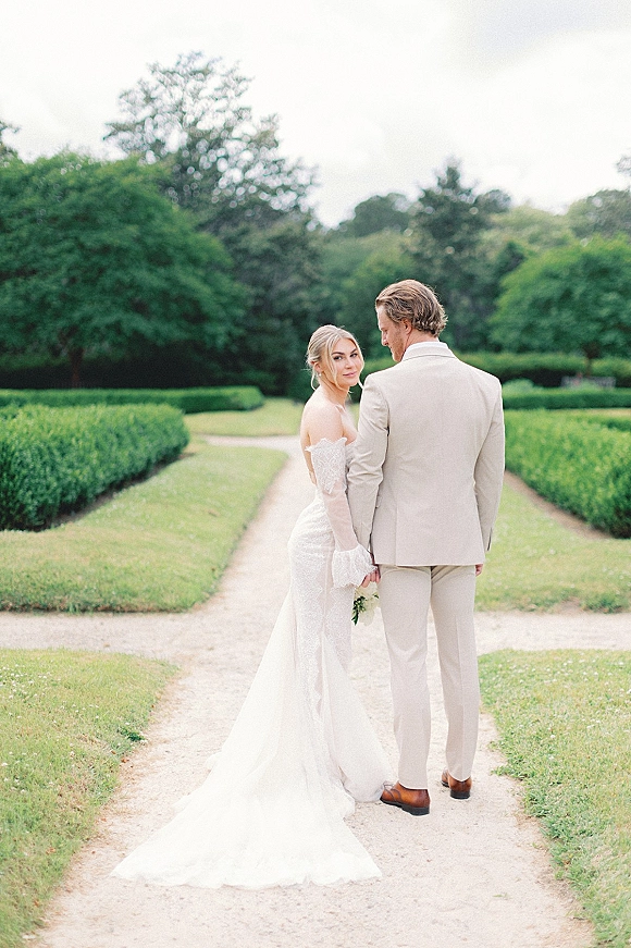 Couple portrait of newlyweds holding hands on a garden path, bride in lace-sleeve gown looking over her shoulder with bouquet