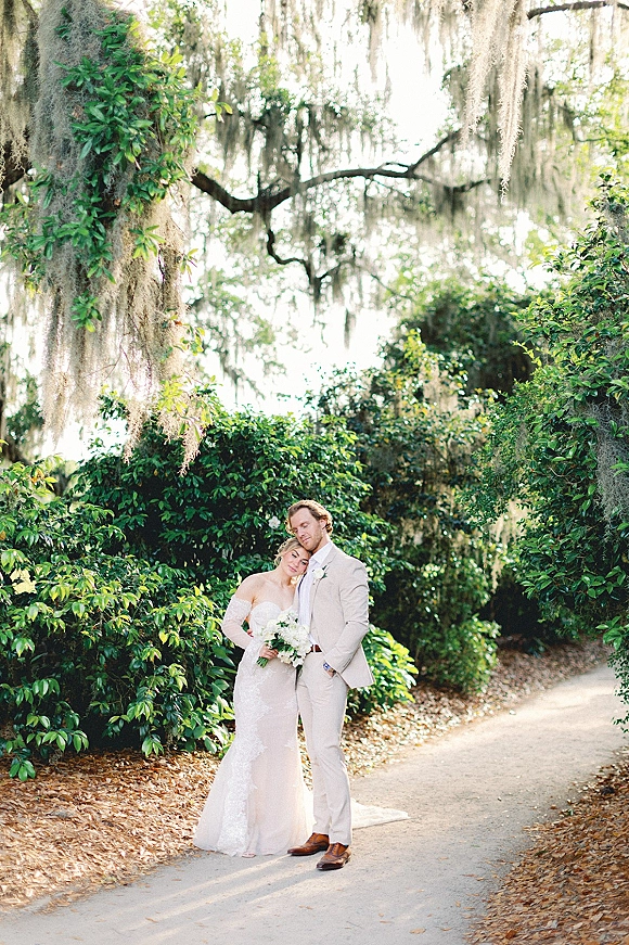 Couple portrait of bride in a strapless lace wedding dress leaning on groom in a beige suit, holding a white bouquet on a garden path under hanging moss