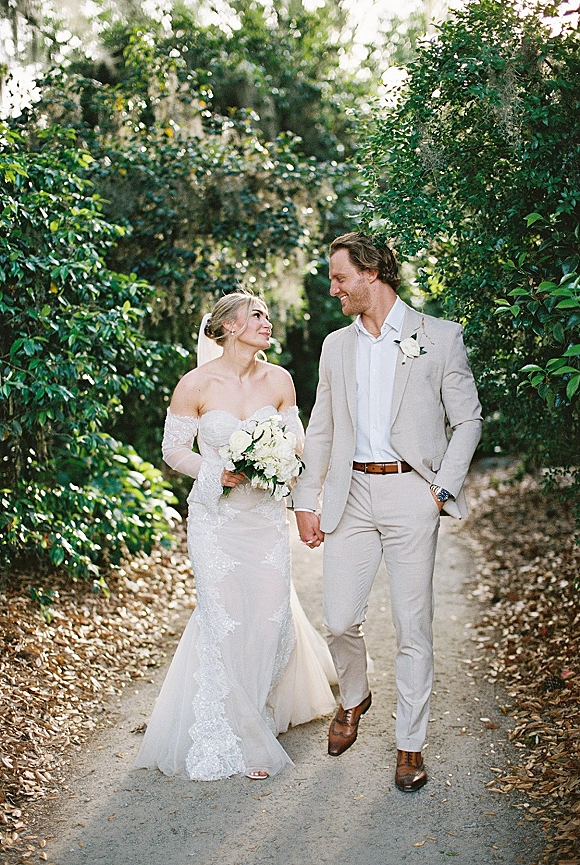 Couple portrait of bride and groom holding hands, walking a tree-lined path, her veil and white bouquet beside his tan suit